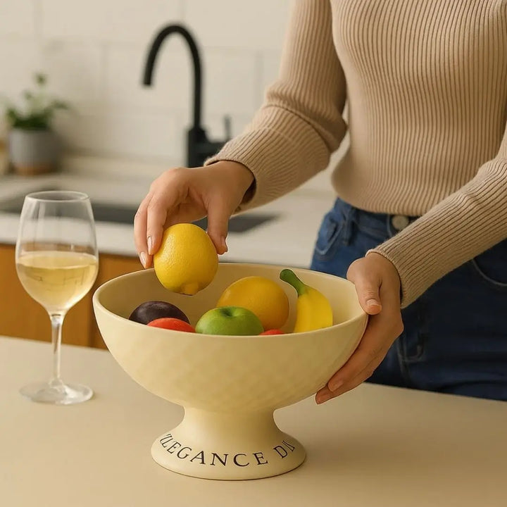 Pessoa organizando frutas na Fruteira Elegance Life Casavelle creme sobre bancada sofisticada com taça ao lado.