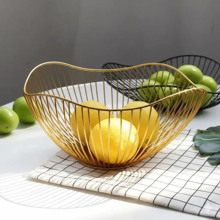 Fruteira Royale Casavelle dourada com limões sobre toalha xadrez em mesa iluminada, com fruteira preta e maçãs verdes ao fundo.