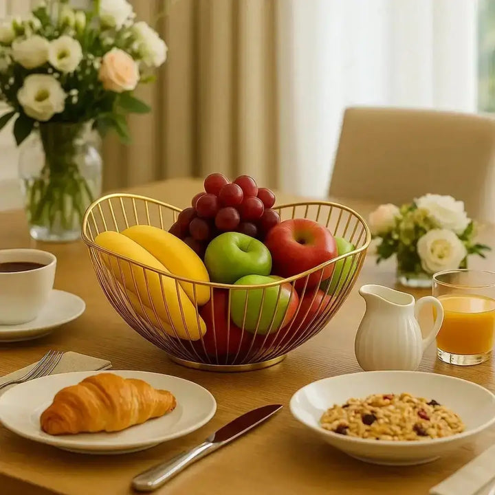Fruteira Royale Casavelle dourada com frutas frescas sobre mesa de café da manhã elegante, ao lado de croissant e taça de suco.