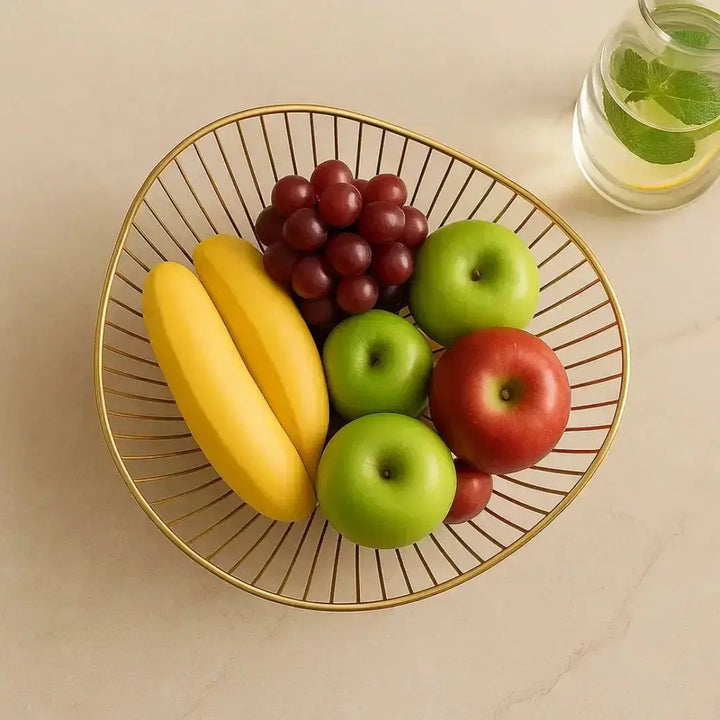 Fruteira Royale Casavelle dourada vista de cima com bananas, maçãs e uvas frescas sobre bancada clara em ambiente iluminado.